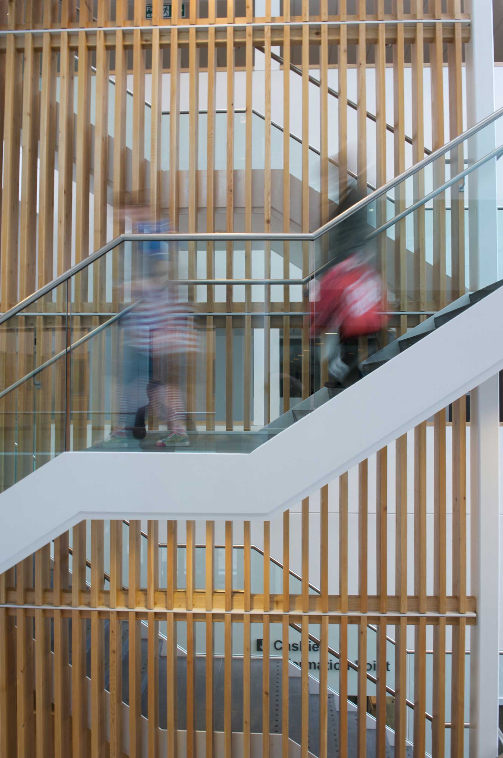 A side on view of a stair case with timber slats and glass sides, with blurred people moving down the stairs 