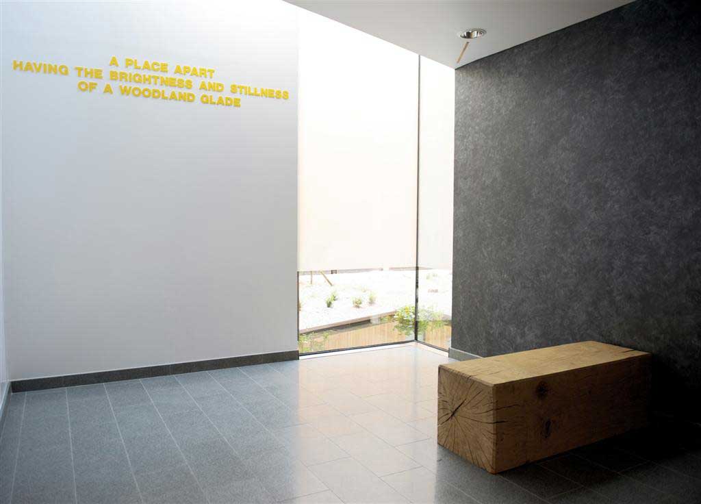 A photograph of a sanctuary space with black wall, a window in the corner and square seat and a white wall with yellow lettering 
