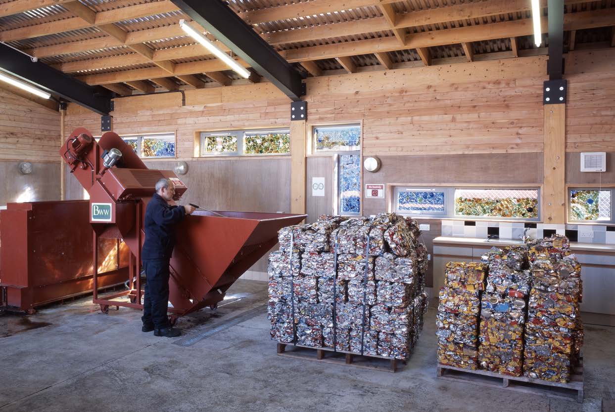 A photograph of a man working in a recycling centre in front of a red machine and two pallets of crushed metal recycling