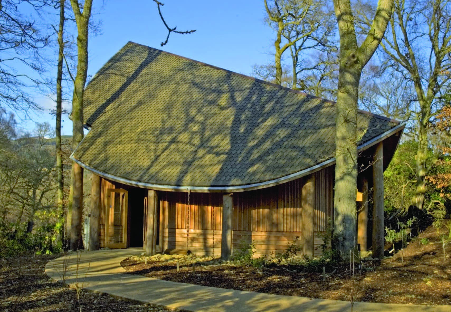 A building with a leaf shaped roof in a forest. A wooden pathway leads up to the building.