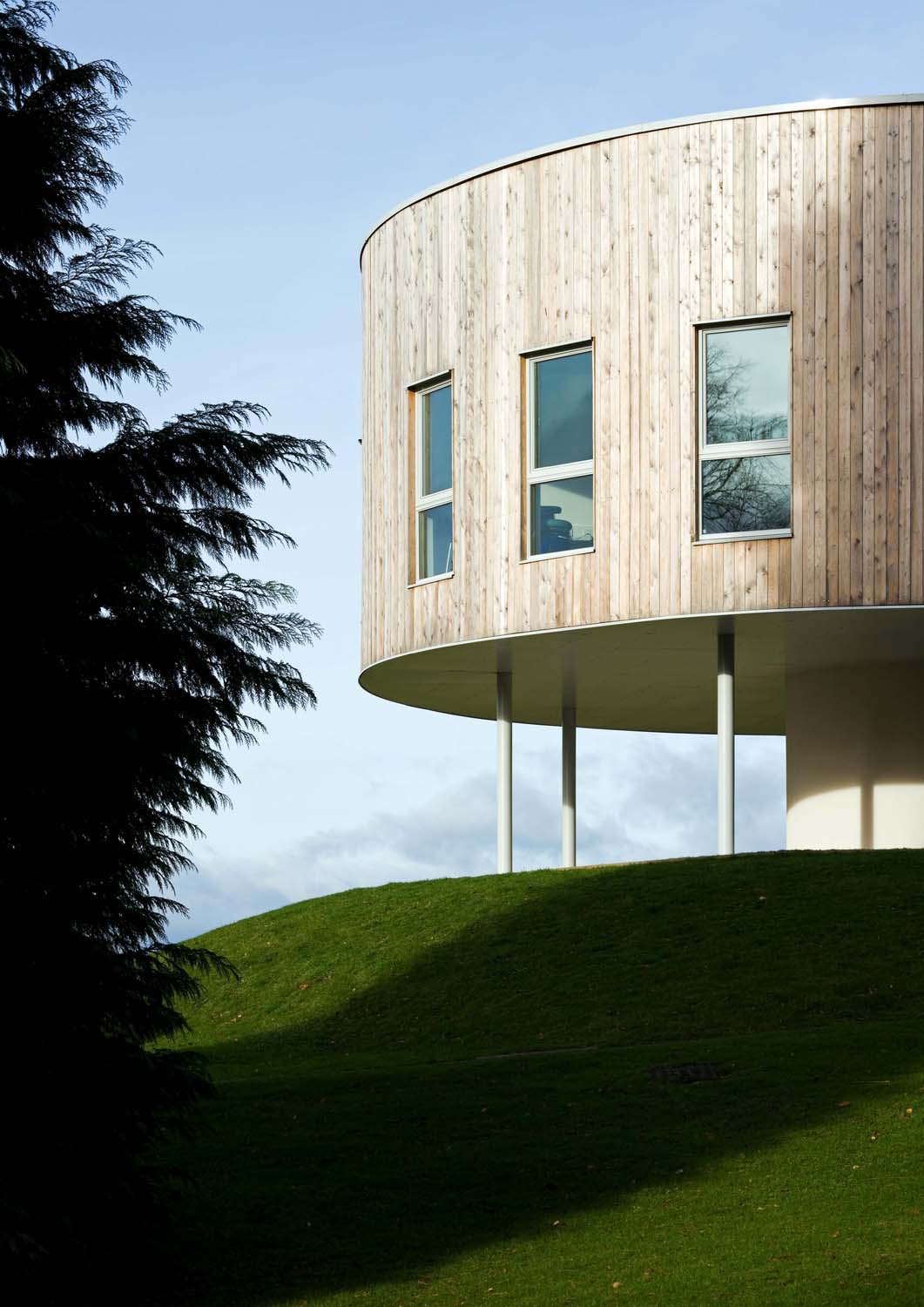 A photograph of a circular timber-clad building on top of a hill, raised from the ground on steel columns 