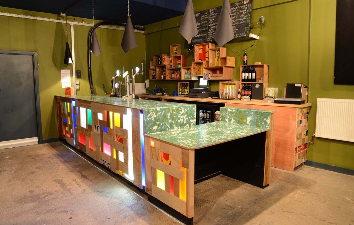 The Glad Cafe bar station's front cover, made out off cut out timber and brightly coloured plastic offcuts. The plastic is lit up by lights.