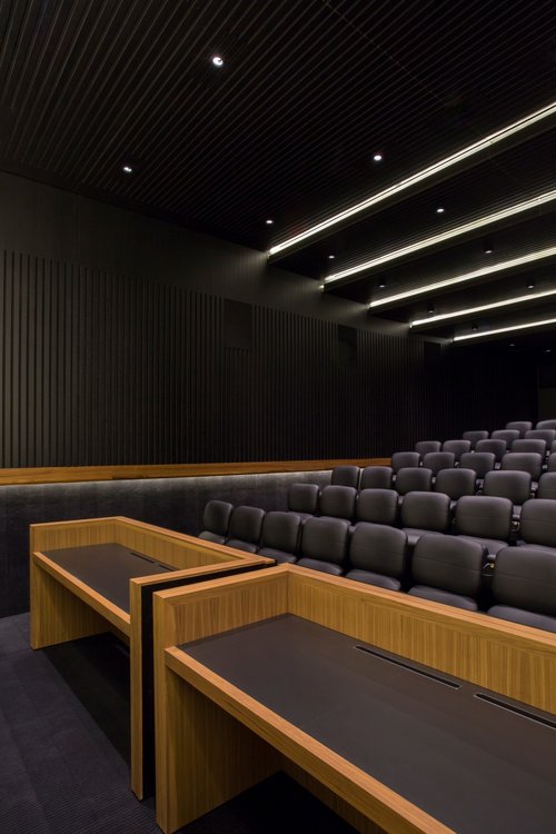 An auditorium with a row of seats. Thee room has dark coloured walls and two desks with a natural wood colour.