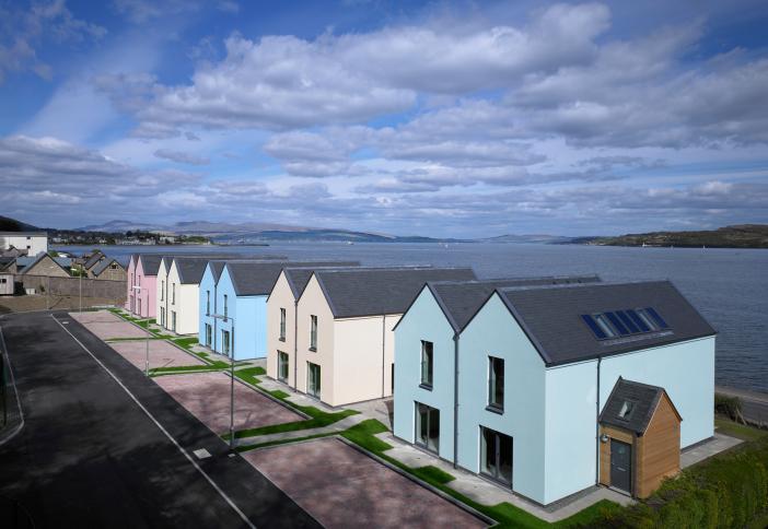An aerial view of a row of different coloured terraced homes overlooking the Firth of Clyde on a partly cloudy day.