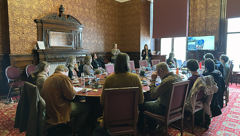 A group of people sit around a table where a presentation is being shown on a scree. The room is dark with has very ornate wallpaper and heavy dark wood furniture