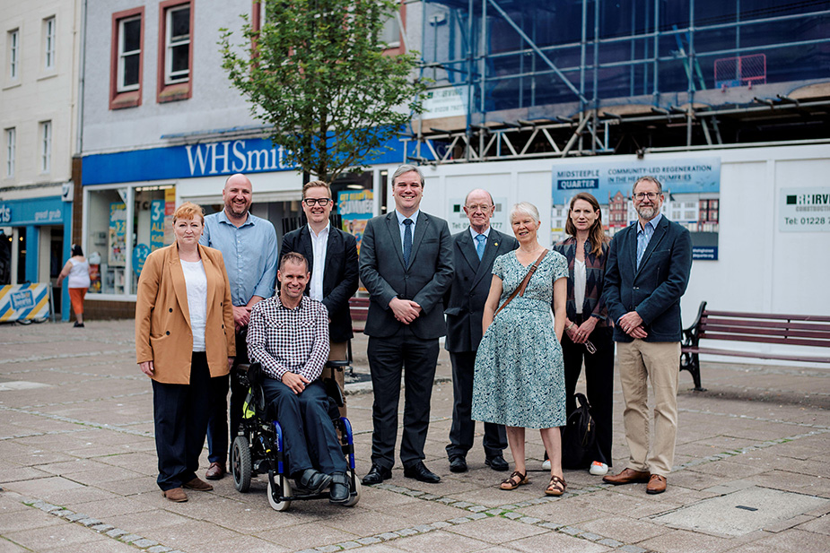 9 people smiling at the camera in front of a building site. There is a man sat in a wheelchair in the forefront.
