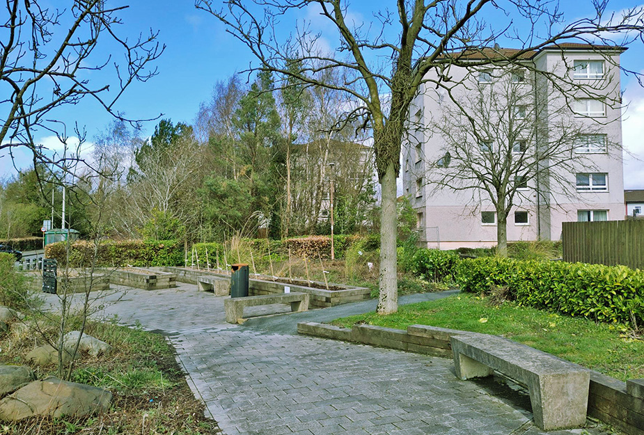 A garden with wide footpath and seating area located nearby a block of flats.
