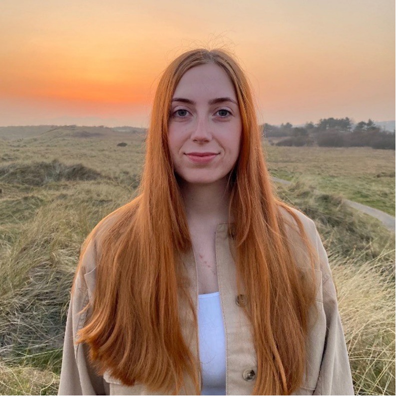 A portrait image of Heather Agnew. She is standing in a field, with an orange sunset in the background. 