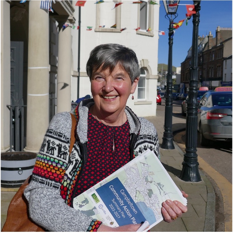 A portrait of Valerie Nimmo .She is standing in front of a stone building and is holding a community action plan document in her hands. 