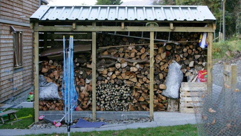 An external wood store filled with wood in various sizes at Kilmun Housing. The structure has a corrugated roof sheet and three walls. 
