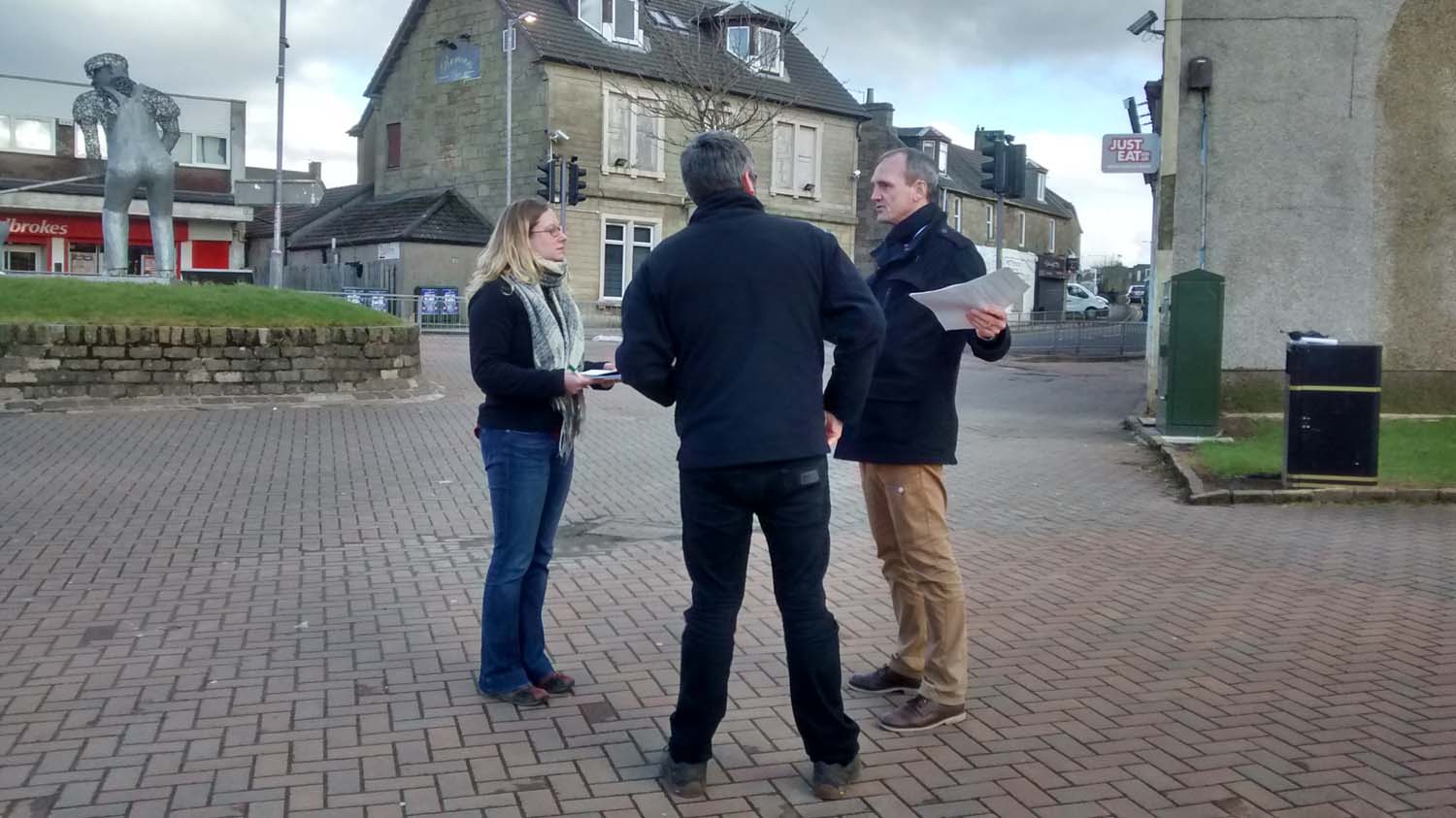 Three people having a conversation about the Place Standard tool beside a building while holding paper.