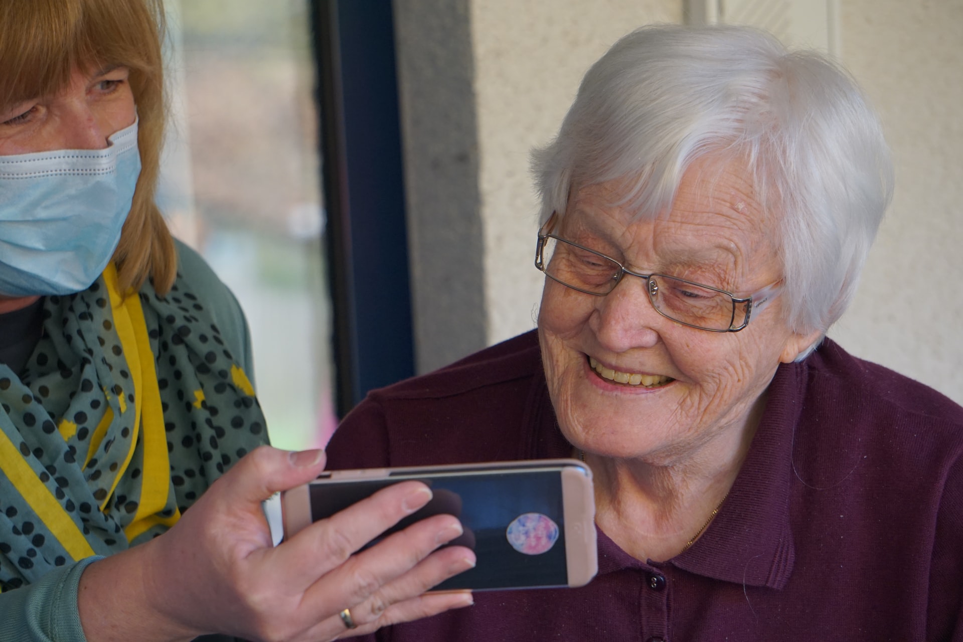 A smiling elderly woman wearing glasses is looking at a smartphone screen.