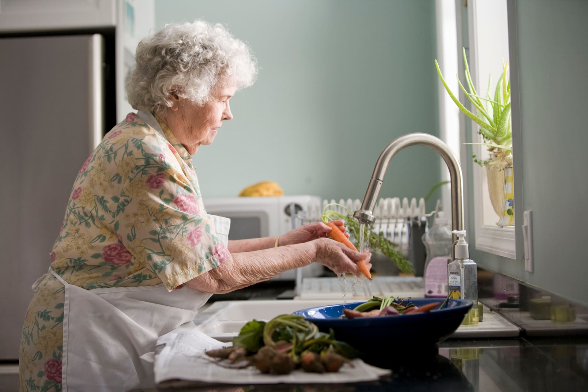 An elderly lady wearing an apron washing carrots in the kitchen sink.