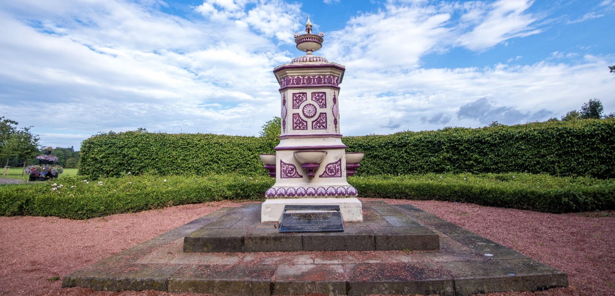 A white and purple monument in a centre of a park. The monument is surrounded by hedges. 
