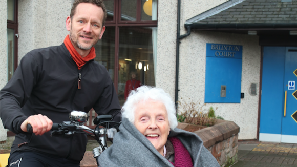 An elderly lady smiling whilst sat on a tricycle in front of a care home.