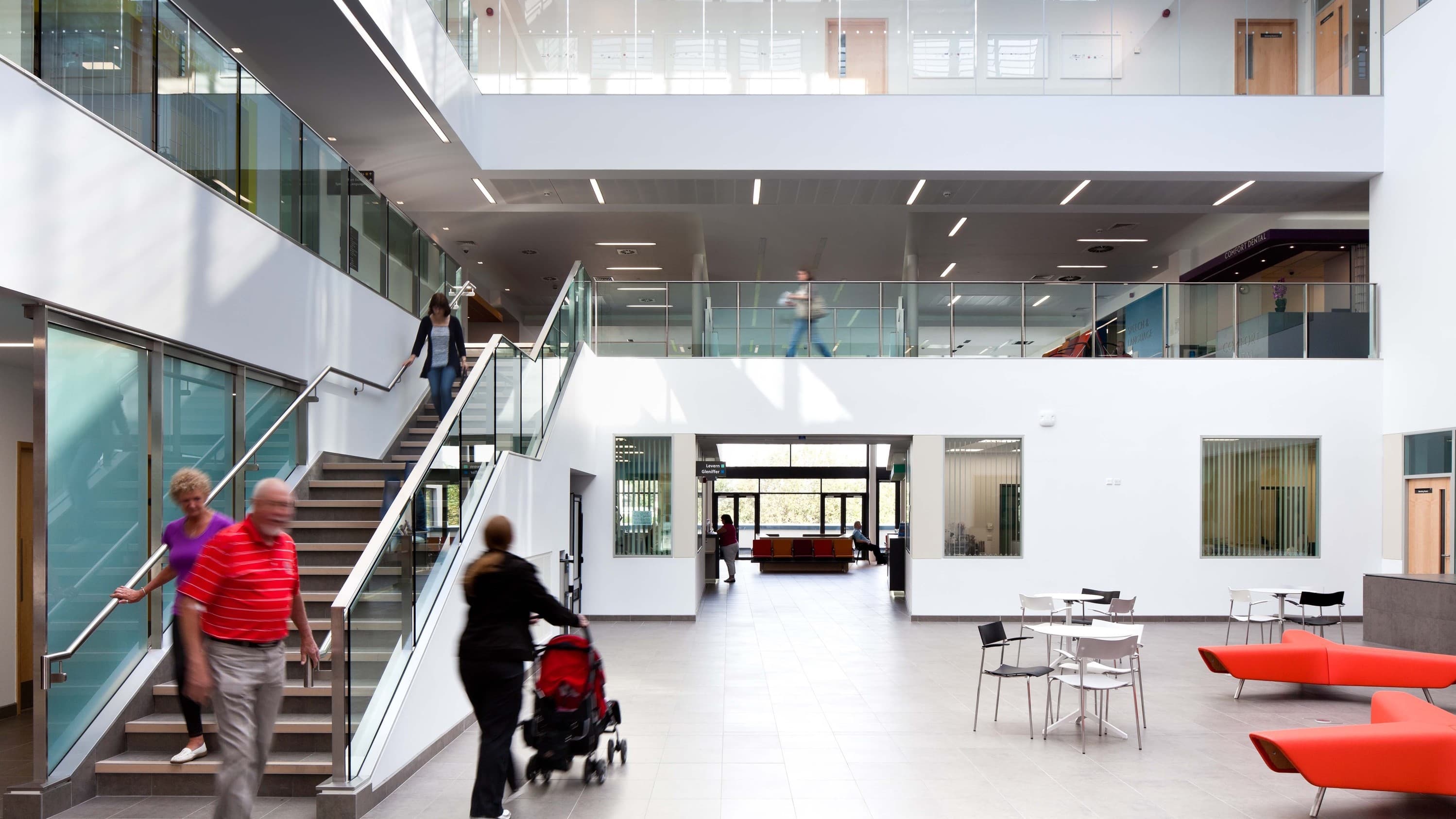 A health centre's double-height main area. There are people walking down a wide staircase on the left side and large windows at the far end of the space.