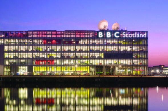 An exterior view of a large modern riverside building at sunset with interior lights reflecting onto the river