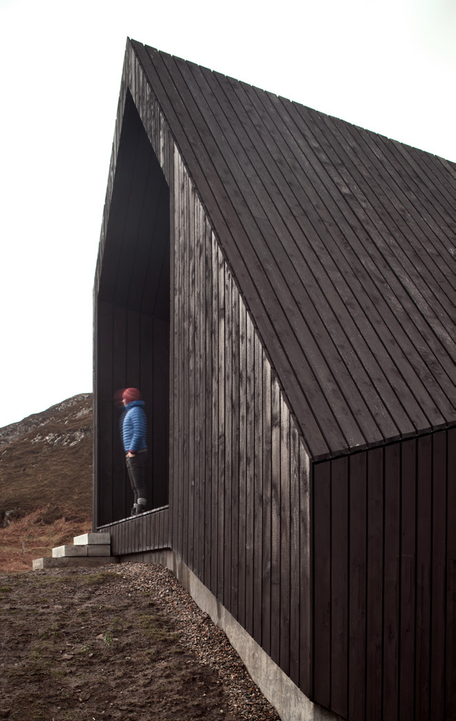 A man standing at the entrance to the House at Camusdarach Sands. Vertical dark wood cladding lines the entire wall and roof of the building.