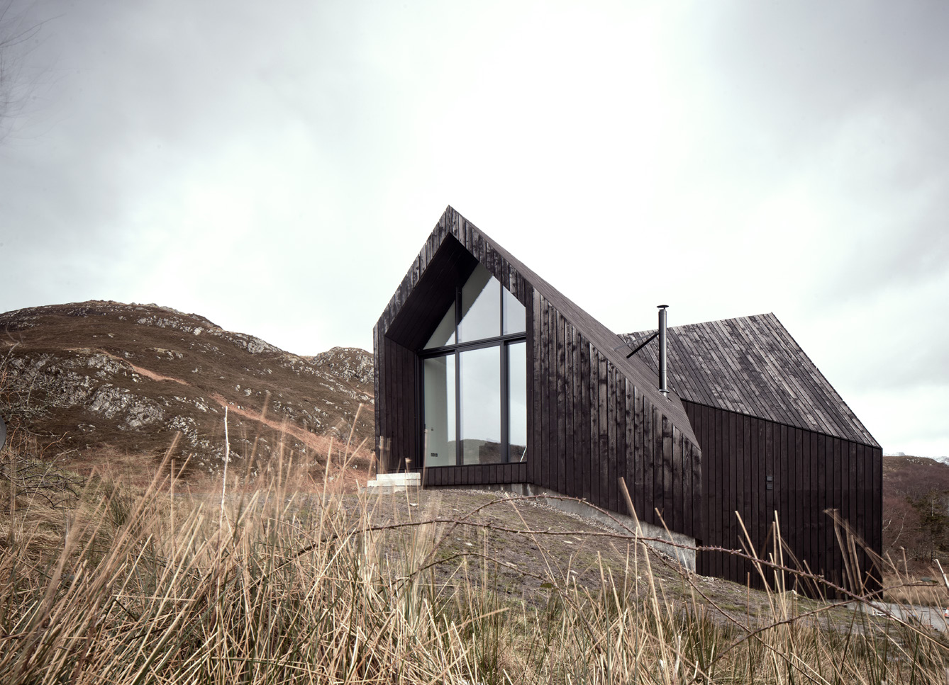 A house at Camusdarach Sands blending into a rural landscape. The house has dark brown timber cladding and with angular shaped roofs.