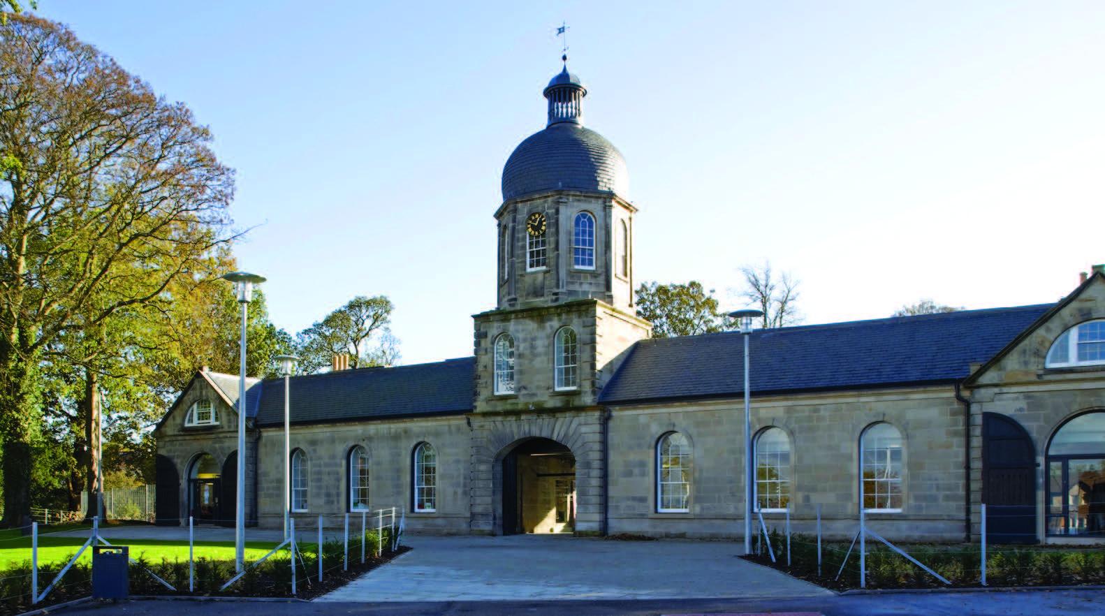 A photograph of castlemilk stables on a sunny day - it is a low rise stone building with an arched entrance and a tower above the entrance