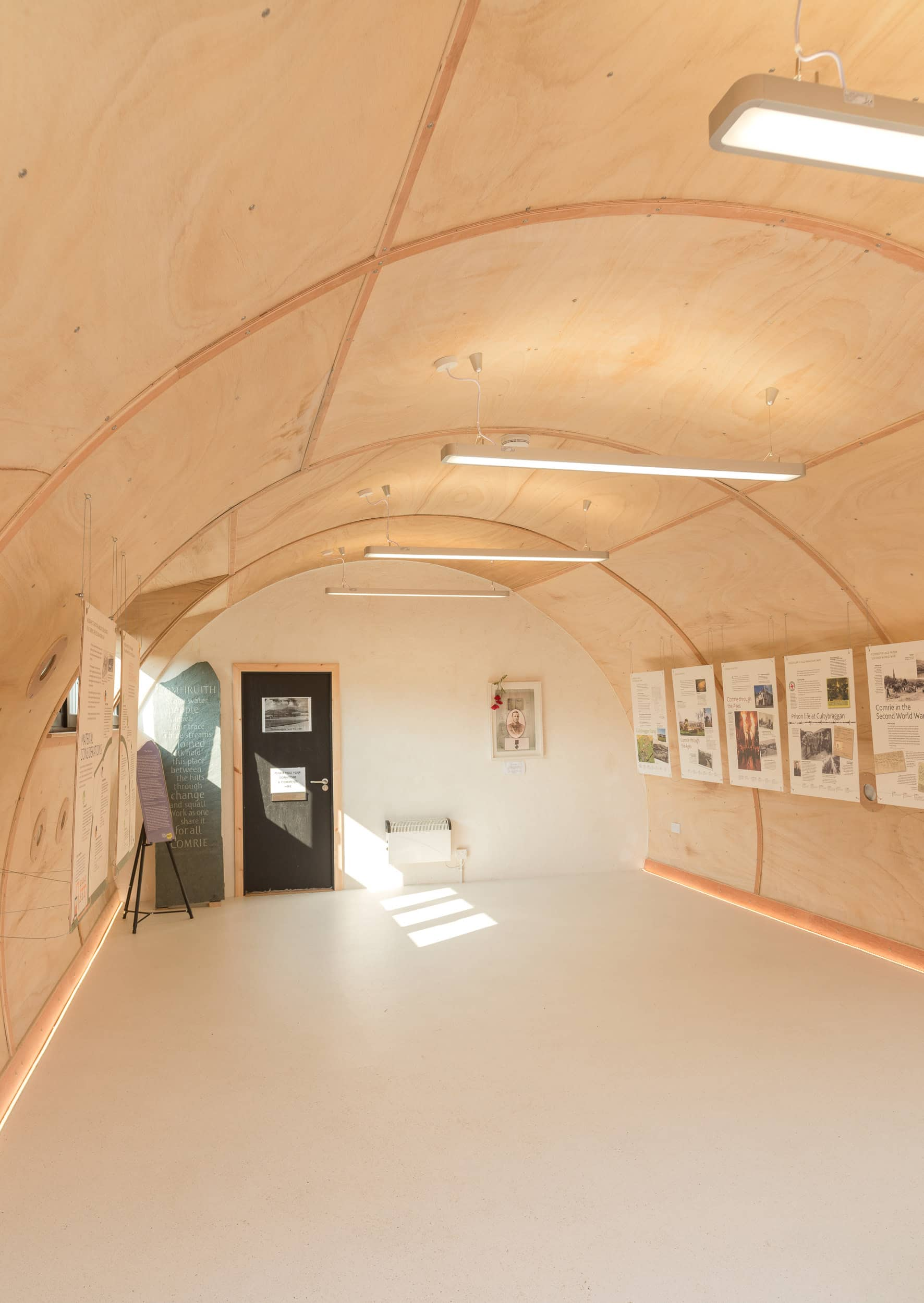 The interior of a narrow and semi circle shaped building. The walls and roof use a pale brown coloured wood with white flooring.