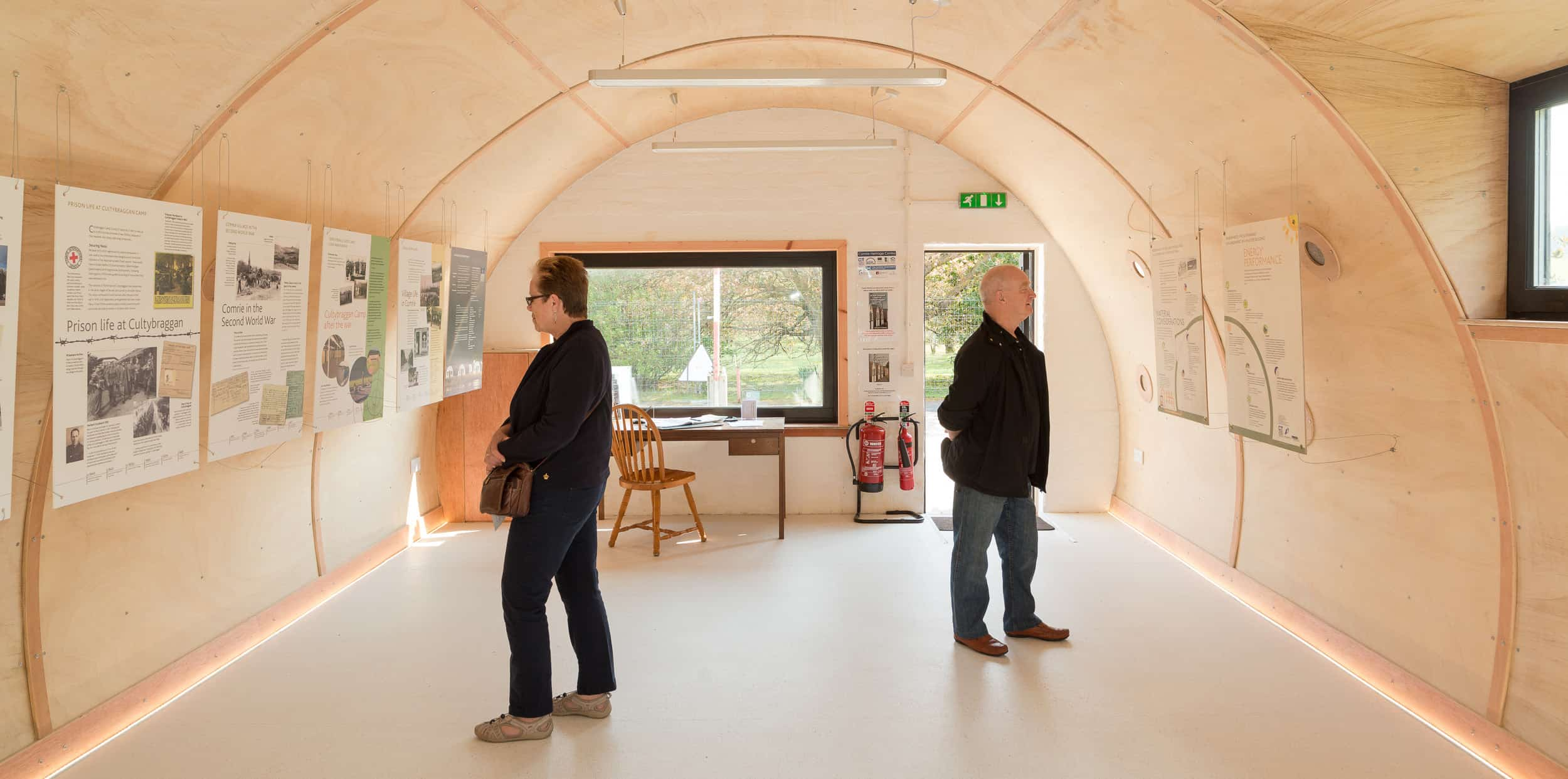 Two people in reading information information located the walls of a semi circle shaped building. The building's interior has a pale colour scheme.