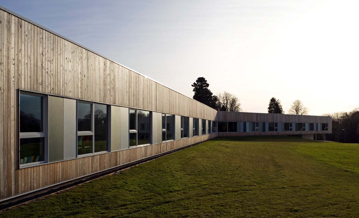 The exterior of a single storey building clad in timber with evenly spaced windows, the sun is setting in the background