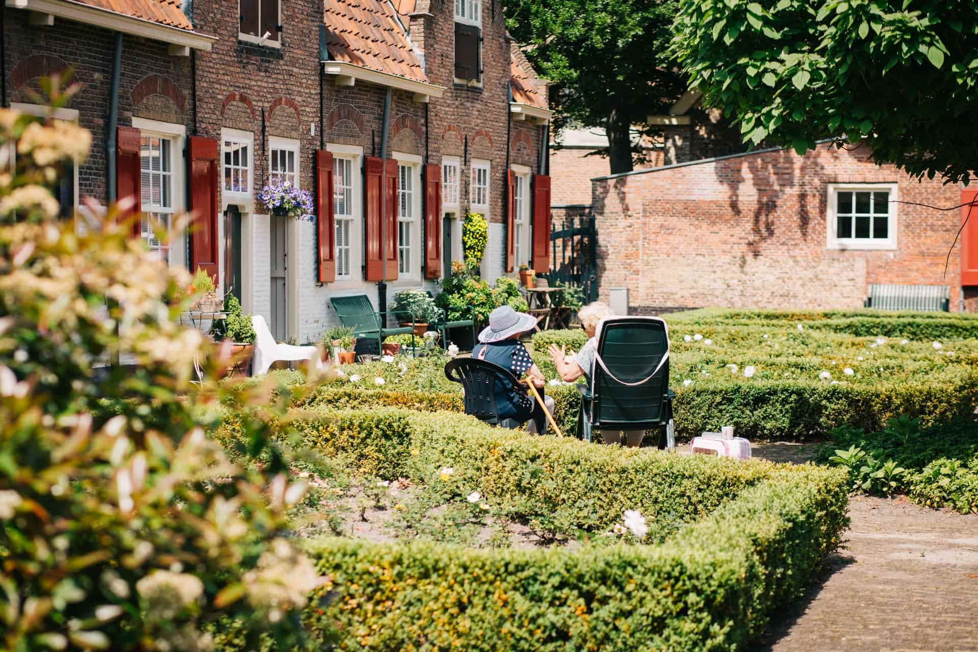 Two elderly people sitting in a garden outside a row of houses with brick facades on a sunny day.