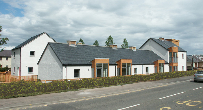 Three attached single-storey houses on the side of the main road with slate roofs. Hedges line the front of the houses.