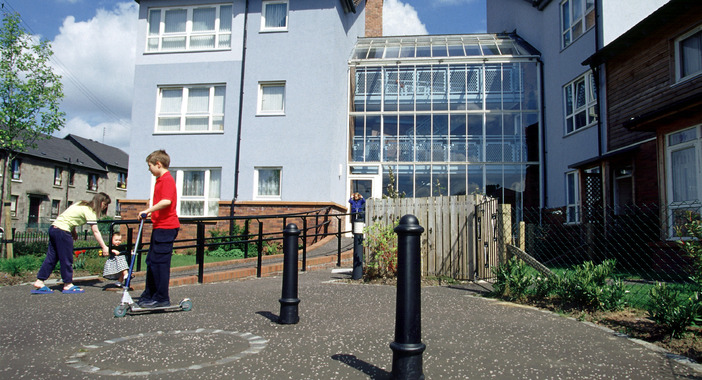 A building with flats and atrium space, with children playing on a pedestrianised street in front of it.