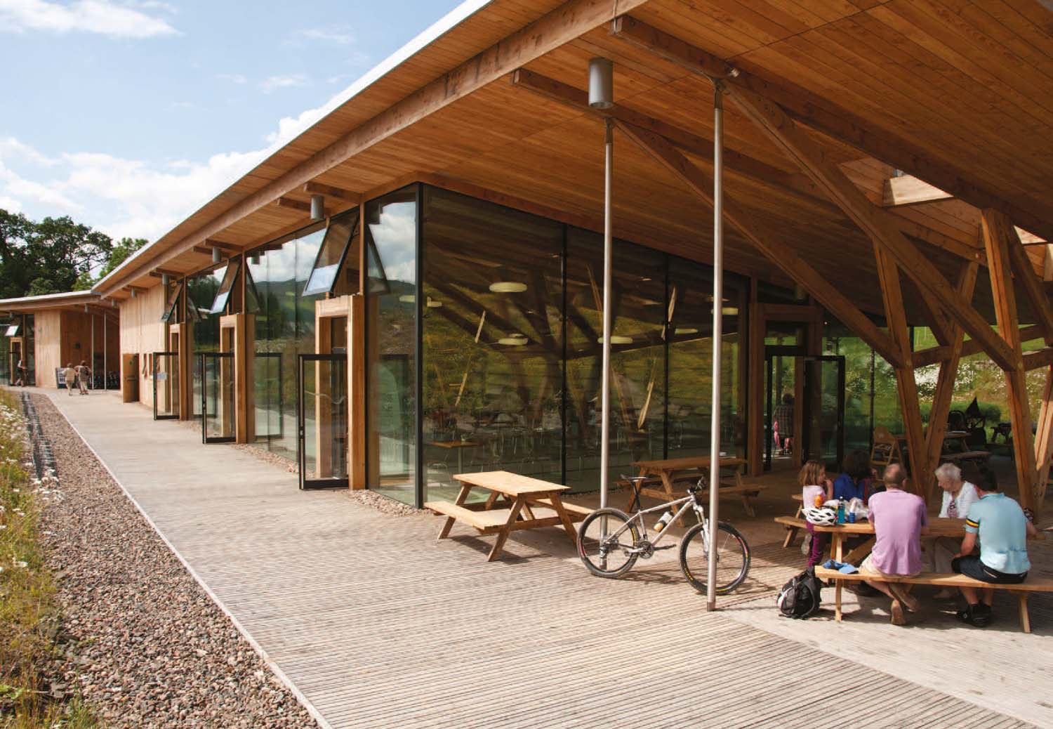 A long and narrow building with timber roof, floor to ceiling glass walls and people sat down in a café with a bicycle.