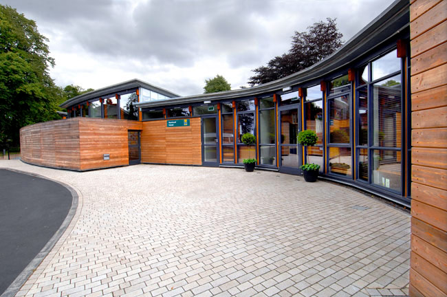 An exterior view of a school building featuring a curved wall with floor to ceiling glass windows and timber elements. Trees are visible behind the school in the background.