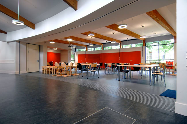 A school's dining area featuring an open plan layout, timber beams and a few brightly red-coloured walls.