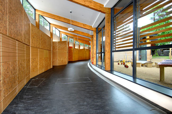 A curved corridor with white ceiling, dark floor and large windows on one side is overlooking a courtyard. The corridor's timber frame is exposed internally.