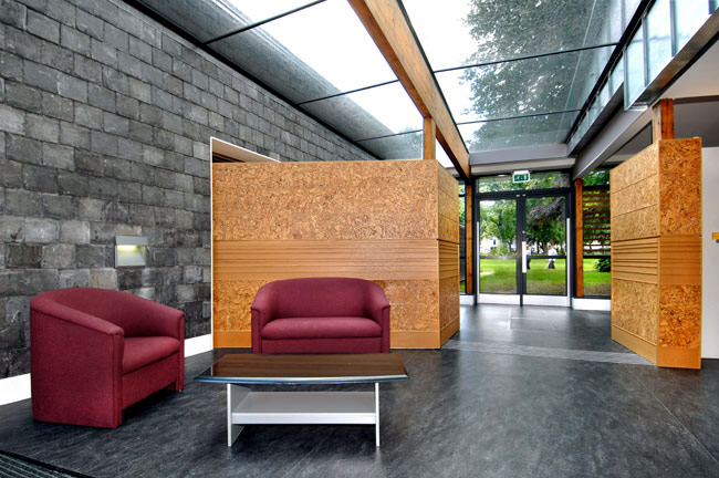 A school's social space positioned under a glass ceiling. There are tactile materials used, such as stone and wood fibre boards. A glass door opposite the space is overlooking a green park.