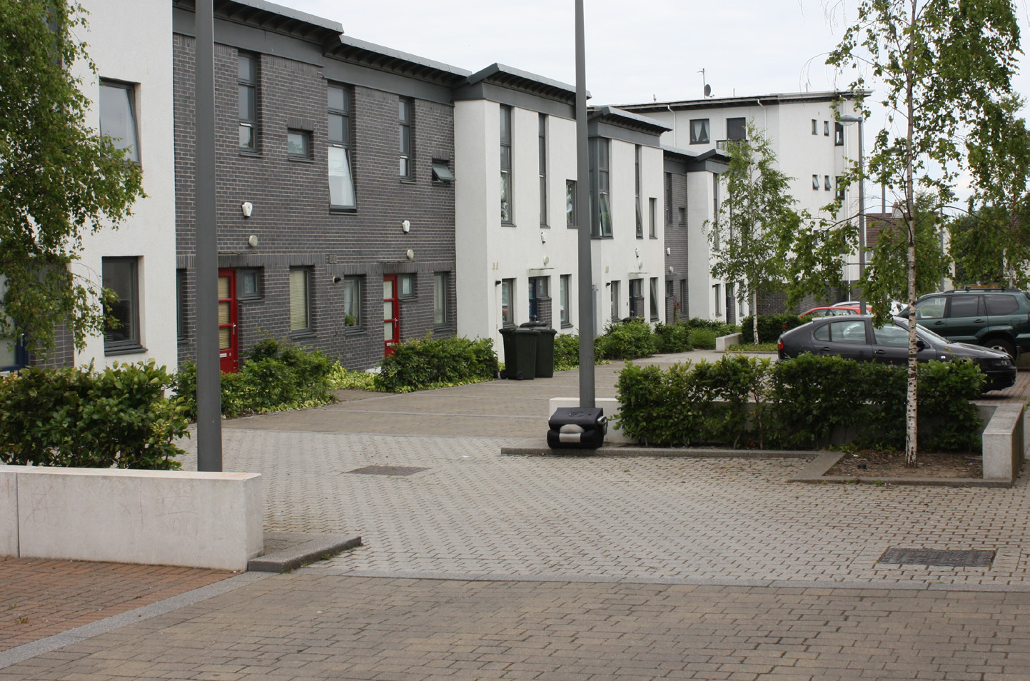 A modern two-storey house mews street with street trees, hedges and four parked cars pictured. The homes are a mixture of white and dark grey.