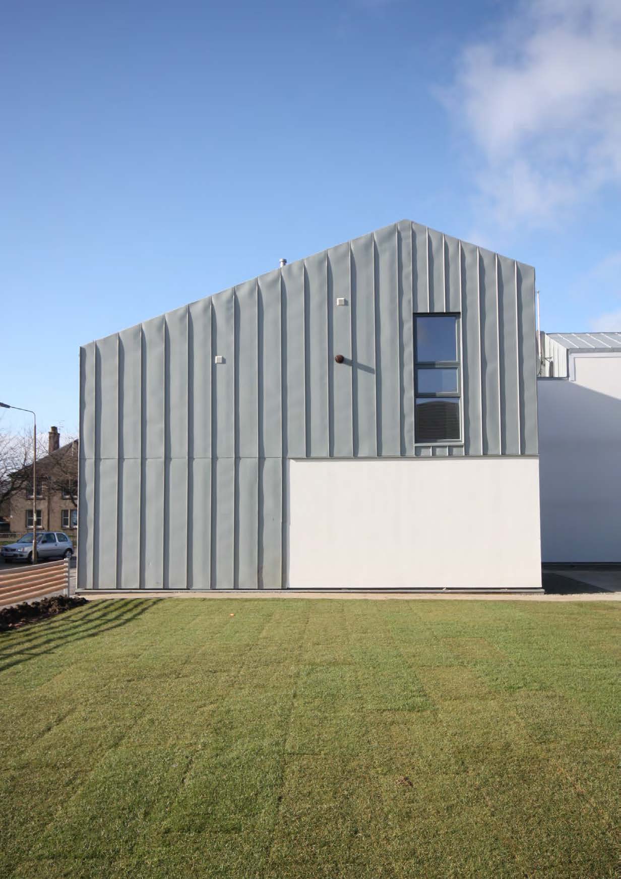 A photograph of the gable end of a two storey building with one tall window, a white rendered wall and grey zinc cladding 