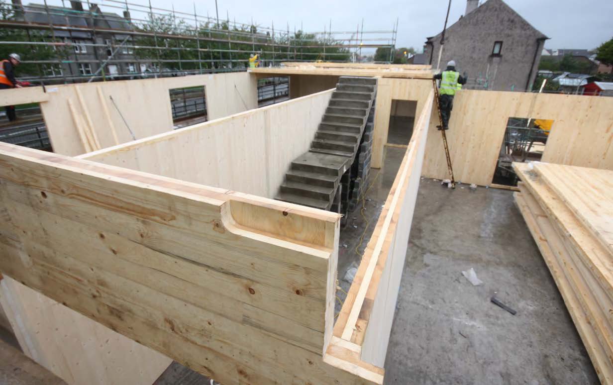 A photograph of the construction of a timber building with the walls and a concrete staircase visible, with a man in a high vis vest and hardhat standing on a ladder