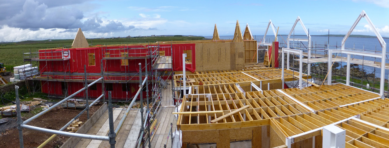 A half-built building of one of the Inns at John O'Groats with scaffolding on-site. The sea is visible on the right and hills on the left. 