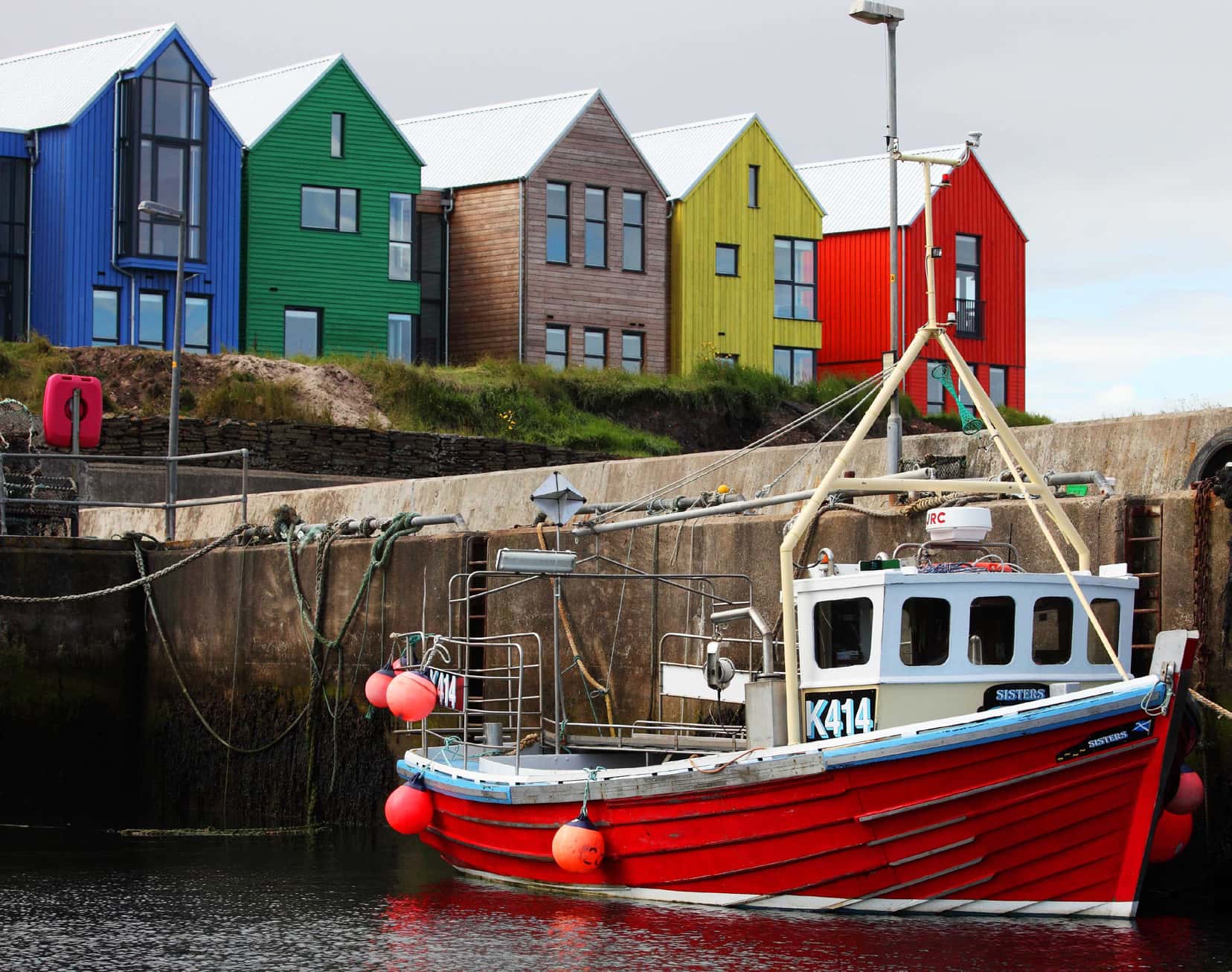 A row of brightly coloured buildings above a stone dock with a bright red boat sitting in the water.