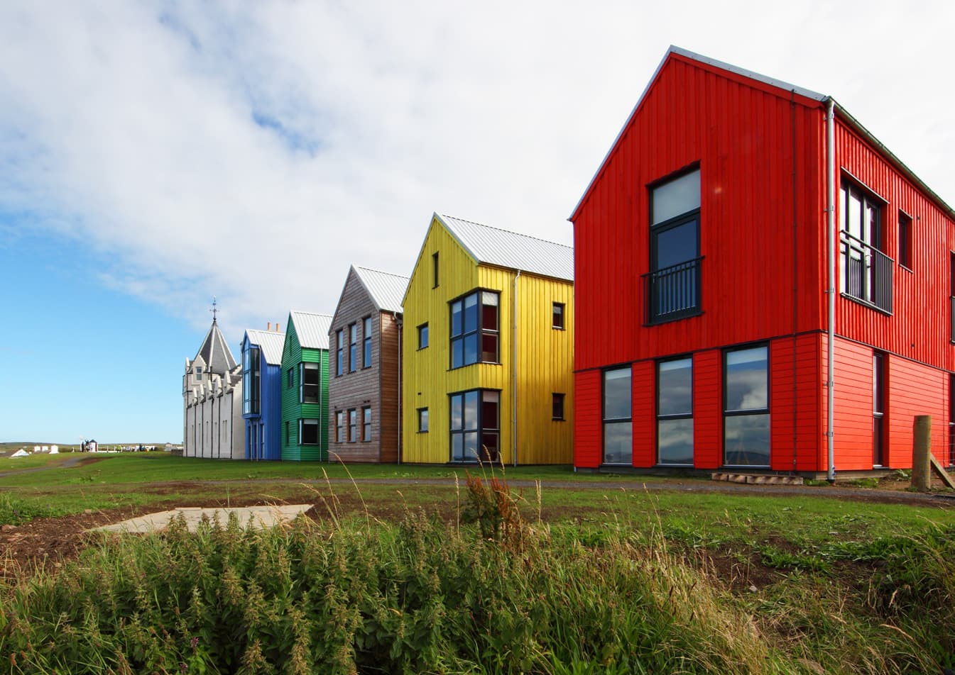 A row of two-storey detached multi coloured building with grass around it. Colours include red, bright yellow, natural wood, bright green, bright blue and white. 