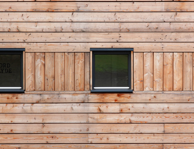 Two square windows on a natural wood coloured external wall made of horizontal wood panels. Vertical pieces are placed beside the windows at the same height.