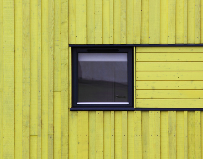 A square window with a black frame surrounded by vertical bright yellow horizontal panels made of wood except for its right side. This is made of vertical panels.