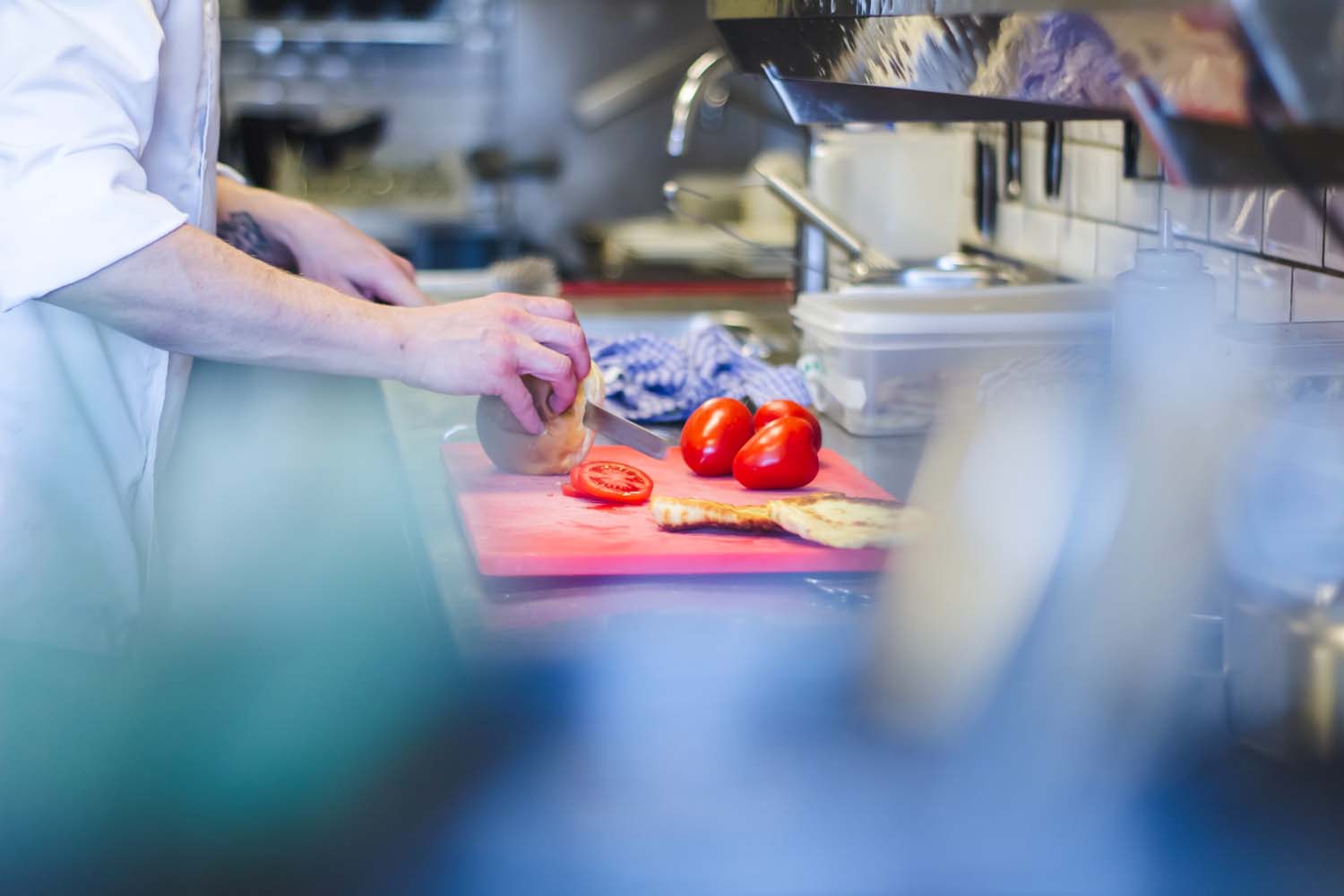 A close up on a pair of hands slicing tomatoes and bread on a cutting board. There is professional kitchen equipment on the background.