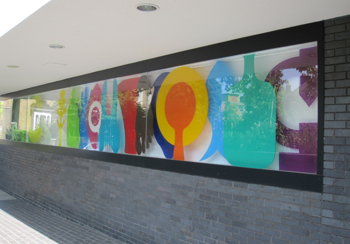 Colourful artwork of different shapes behind glass stretching across the length of a grey outside wall.