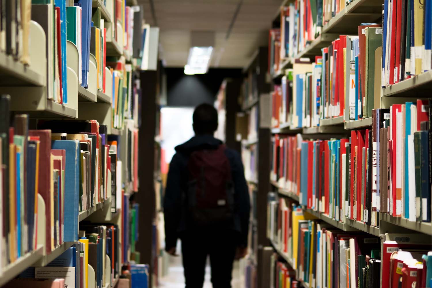 A photograph of a person walking away from the camera between two full bookshelves of colourful library books
