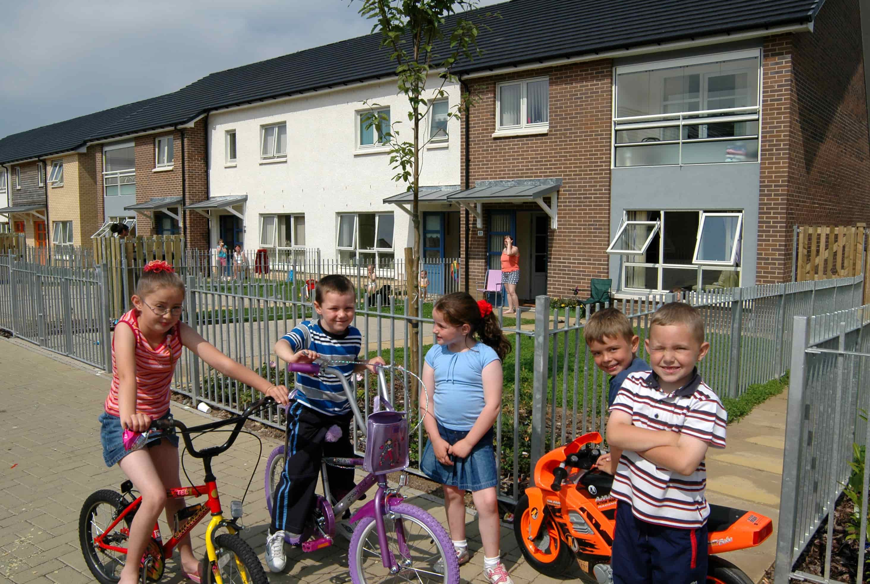 A group of smiling children posing with their bicycles on the pavement in front of their houses.