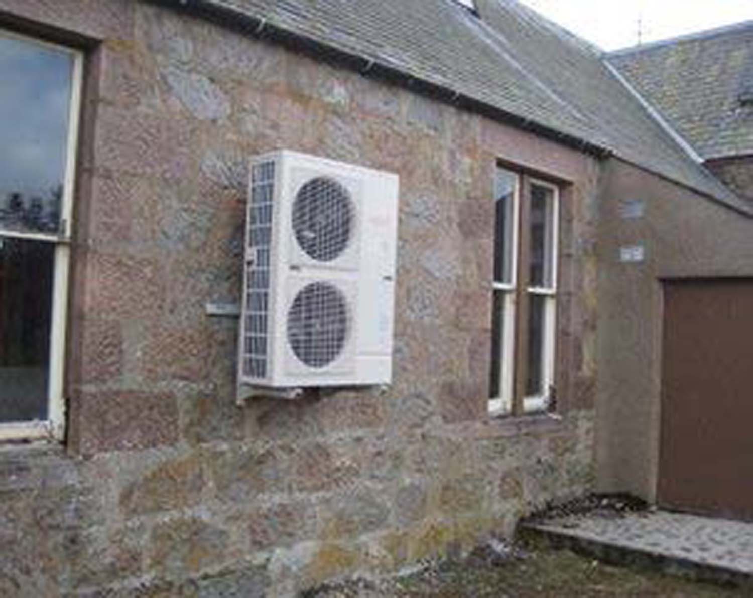 A photograph of a white air source heat pump on the grey stonewall of a single-storey building