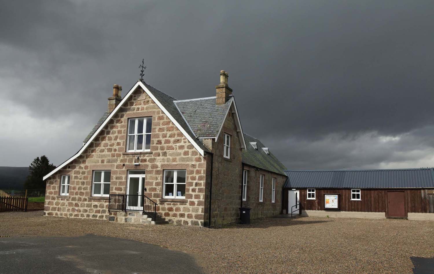 The exterior of a light grey stone building in the countryside with a dark grey sky above