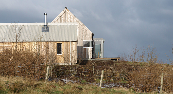 A side view of the Long House which is set in a rural setting. The house's exterior timber is a faded silver grey paired with a grey corrugated roof.